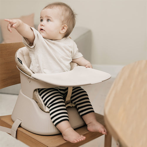 Baby sitting in a beige baby seat on a wooden dining chair.